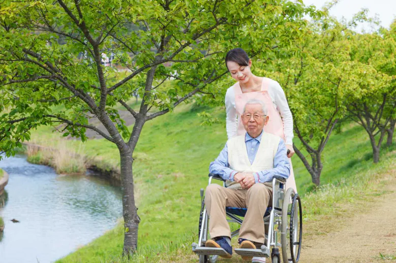 Caregiver pushing an older man in a wheelchair along a path by a stream
