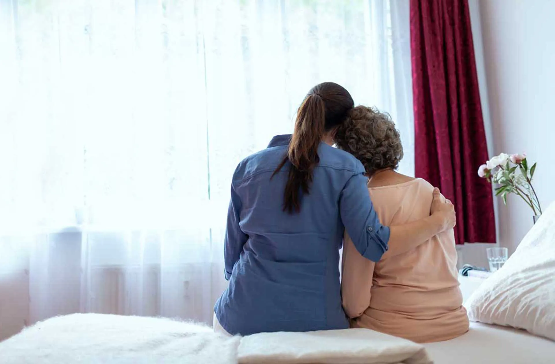 Younger woman with her arm around an older woman as they sit on a bed facing a window