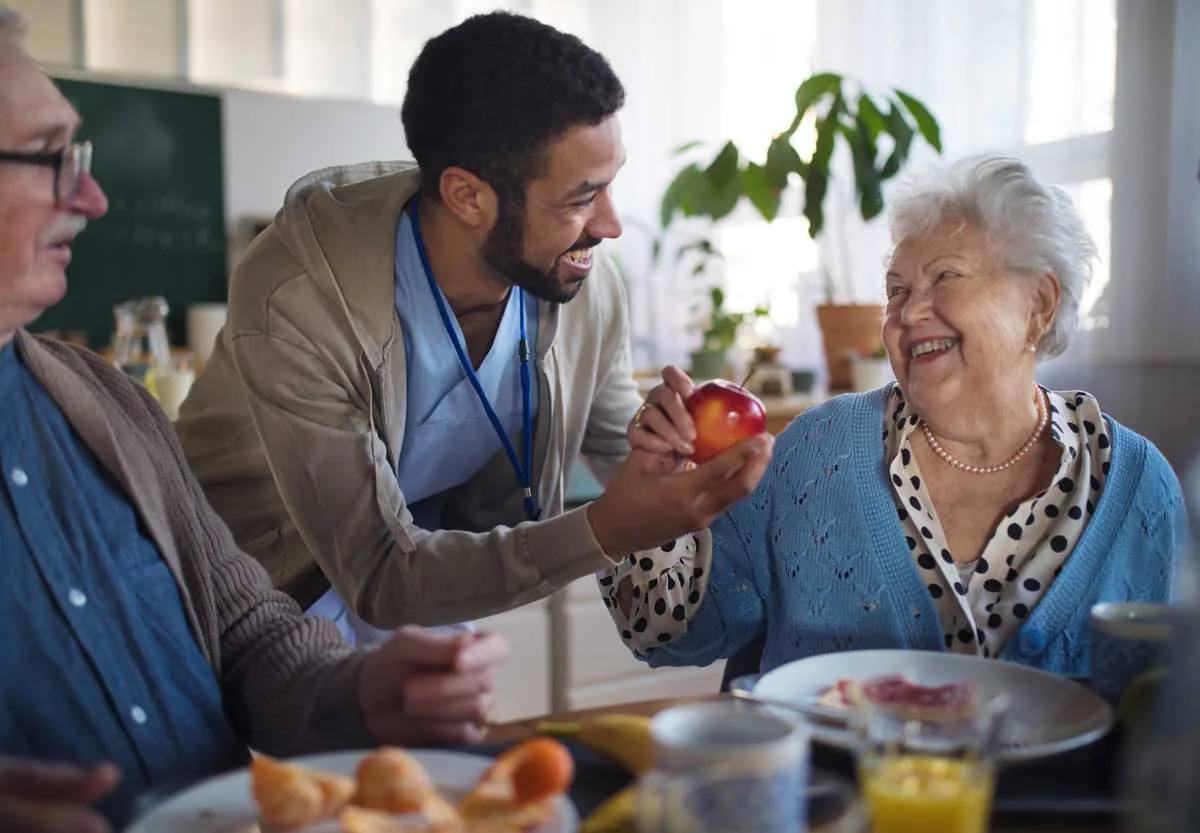 Caregiver handing an apple to a smiling older woman at breakfast