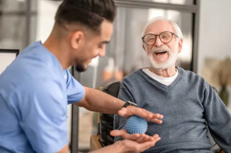 Caregiver in scrubs showing a massage ball to a laughing older man