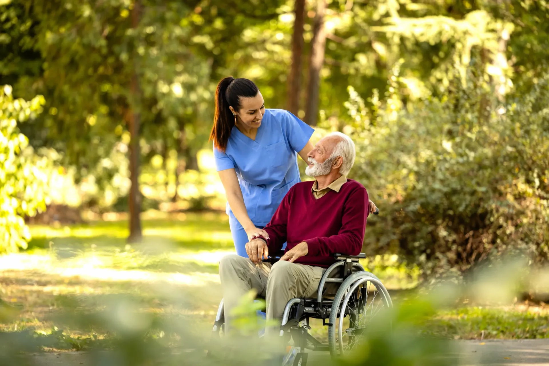 Caregiver in scrubs with a smiling older man in a wheelchair outdoors